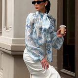 Elegante blusa con mangas farol y cuello atado, adornada con flores.
