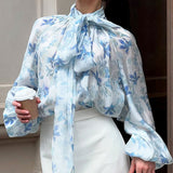 Elegante blusa con mangas farol y cuello atado, adornada con flores.
